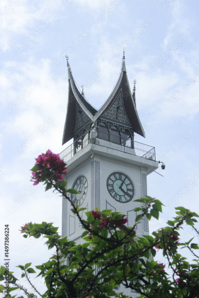 Fototapeta premium A close-up of the iconic Jam Gadang clock tower in Bukittinggi, Indonesia, featuring traditional Minangkabau architecture. Pink flowers in the foreground add a vibrant touch to the elegant structure u