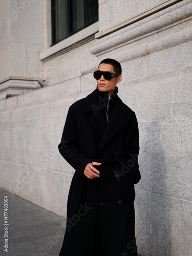 Portrait of handsome Chinese young man wearing black overcoat posing in the street, young guy with black short hair all in black with urban background. Male fashion, cool Asian young man lifestyle.