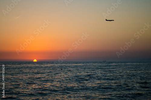 Foto Airplane glides above calm ocean, silhouetted against vibrant sunset