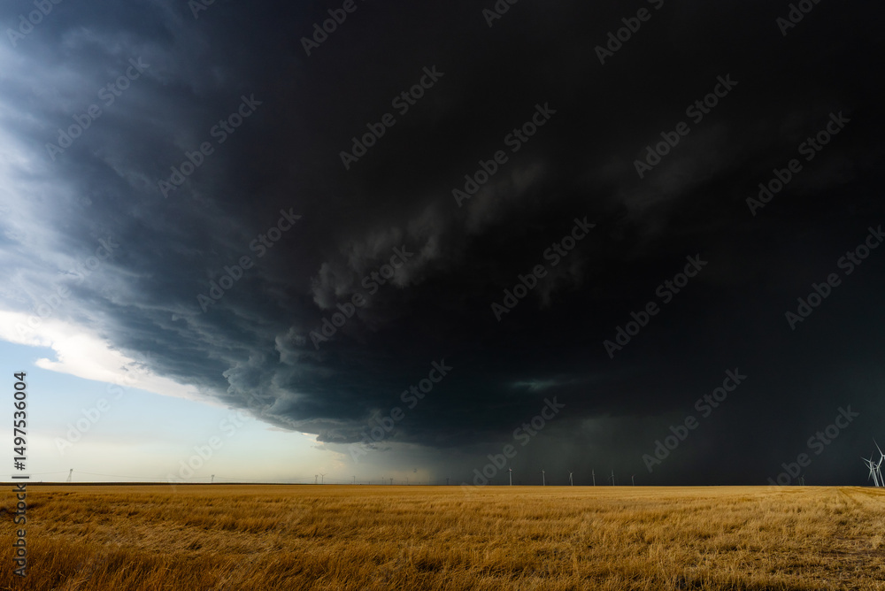 Fototapeta premium Storm clouds roll across the sky above warm-colored fields and a wind farm, creating a dramatic, moody scene filled with texture, motion, atmosphere, and the beauty of nature and weather.