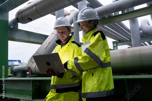 Engineers collaborate on a project at an industrial site during daylight hours wearing protective gear and using a laptop
