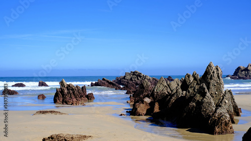 Dramatic jagged rock formations on a sandy beach in Asturias, Spain with gentle ocean waves in the background.