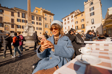 Woman in a blue coat scrolls her phone with a smile at a café table in a vibrant Roman square, bathed in warm sunlight