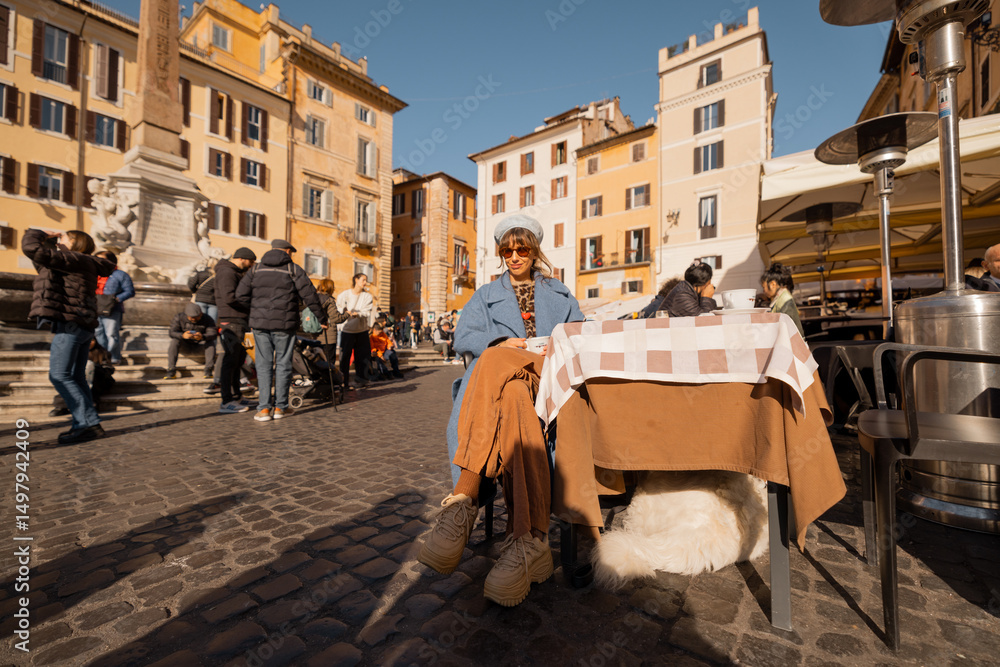 Obraz premium Smiling woman in a blue coat enjoys her coffee in a sunlit Roman square, surrounded by warm-toned buildings and people strolling by