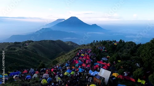 A vast mountain range with a dense campsite at the foot of a peak, shrouded in fog under a pale blue sky