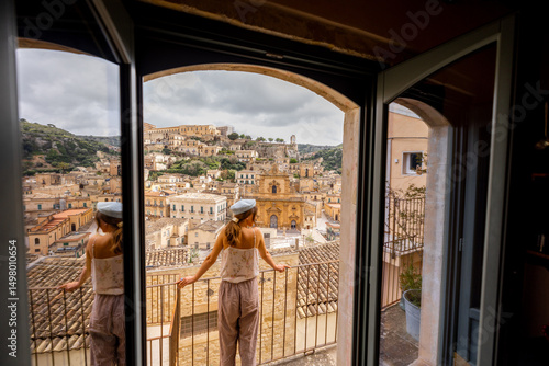 Woman standing on a balcony with panoramic view over the baroque rooftops and historic church of a hilltop Sicilian town. Reflection in glass door, soft light and serene atmosphere