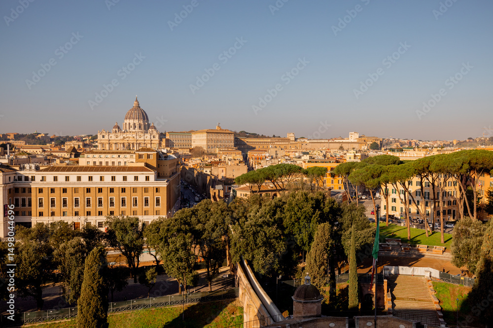 Fototapeta premium Panoramic view of Vatican City and the dome of St. Peters Basilica from the top of Castel Sant Angelo, framed by Roman pines and morning sunlight over the rooftops of Rome