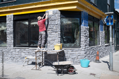 Construction worker standing on a tressel fixing stone facing to an exterior wall around a window on the street.