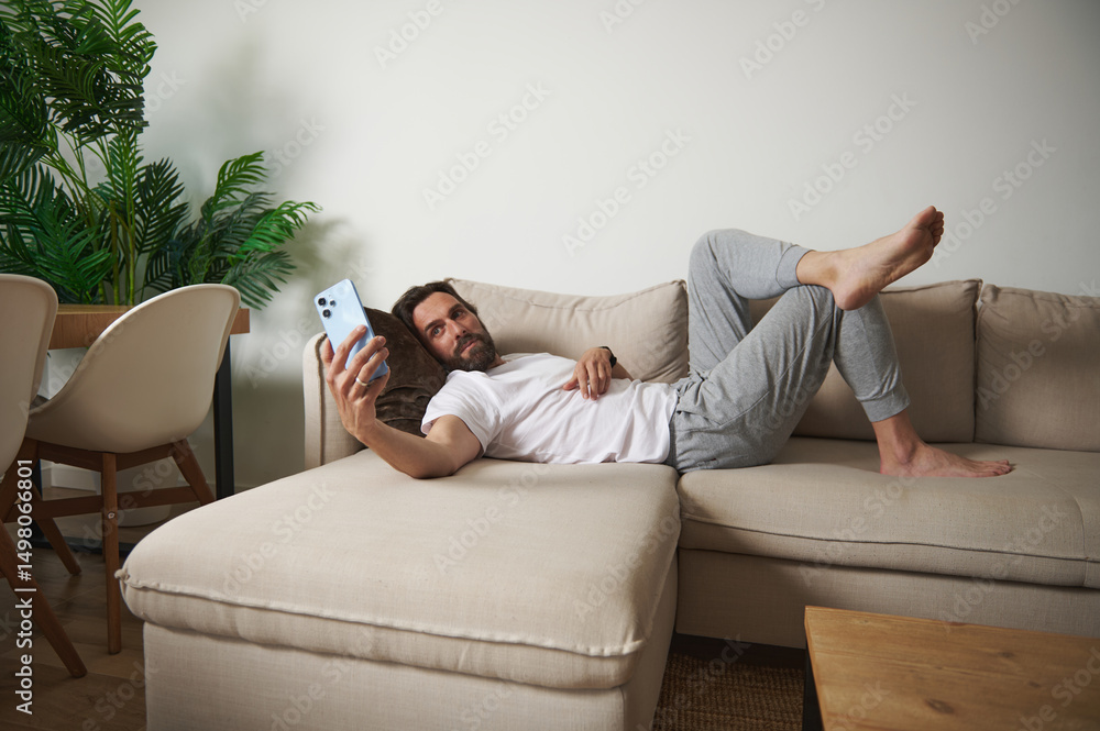 Obraz premium Man Relaxing on Sofa Holding Smartphone in Cozy Minimalist Living Room Setting