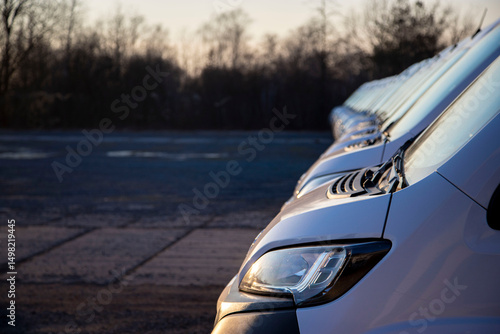 Side and front of a panel van in a long row of identical white vans ready for customer delivery. Headlight, hood, fender, bumper. A line of vans in a logistics parking lot.