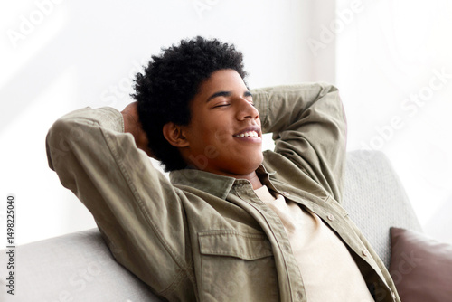 Happy black teen guy relaxing on comfortable sofa at home during coronavirus quarantine. Positive African American teenager resting on couch, spending lazy time on weekend morning