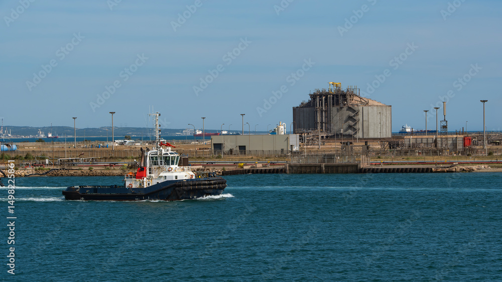 A tugboat moves past a large, concrete LNG storage tank in a busy industrial port A tugboat moves past a large, concrete LNG storage tank in a busy industrial port