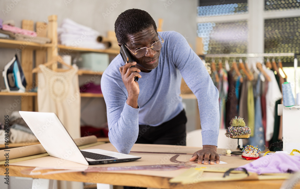 Experienced African American male tailor communicating on phone with client or supplier while reviewing specifications and timelines on laptop in sewing workshop Experienced African American male tailor communicating on phone with client or supplier while reviewing specifications and timelines on laptop in sewing workshop