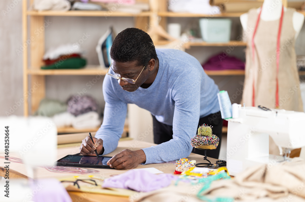 Fototapeta premium Dedicated male tailor concentrating on designing custom dress using tablet, reviewing and adjusting clothing model in well-equipped creative studio