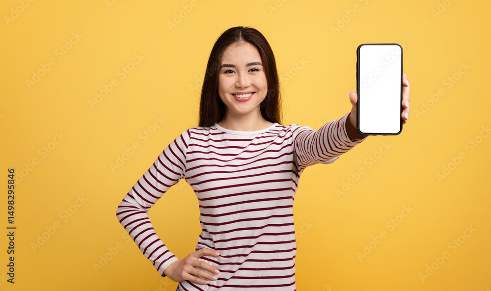 Naklejka premium Asian young woman with long dark hair is smiling while holding a smartphone in front of her. The vibrant yellow background enhances her cheerful demeanor and the blank screen invites curiosity.
