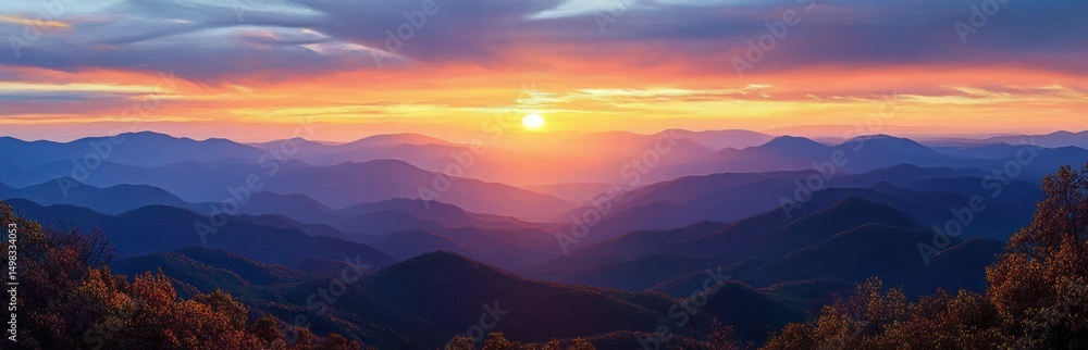Obraz premium Sunset over a vast mountain range with layered ridges and colorful sky casting warm light and shadows on foliage in the foreground