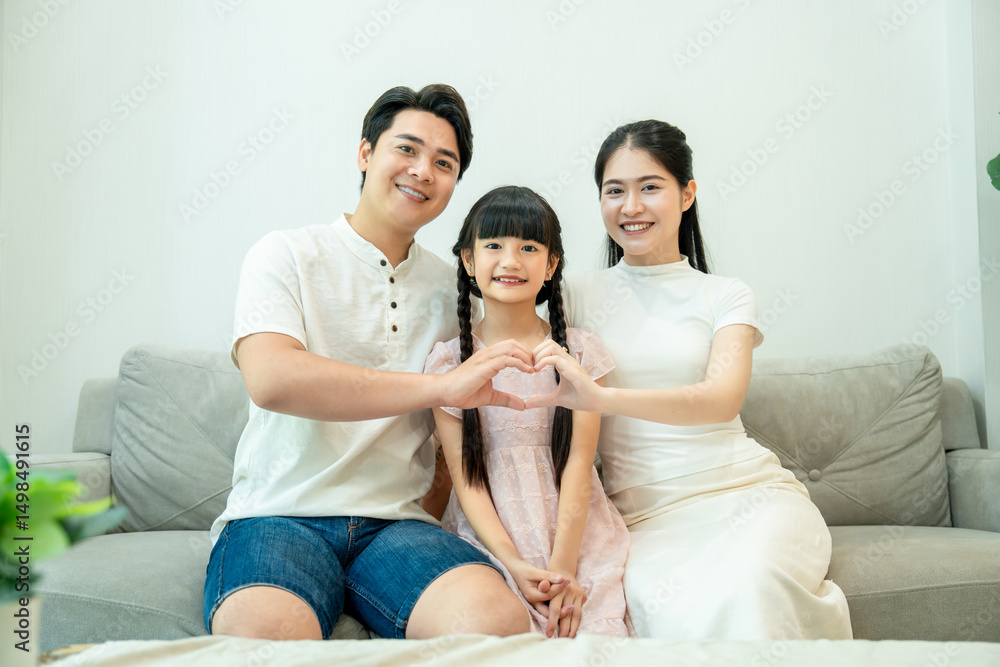 A loving Asian family is sitting on a couch. The parents are making a heart shape with their hands in front of their smiling daughter. A loving Asian family is sitting on a couch. The parents are making a heart shape with their hands in front of their smiling daughter.