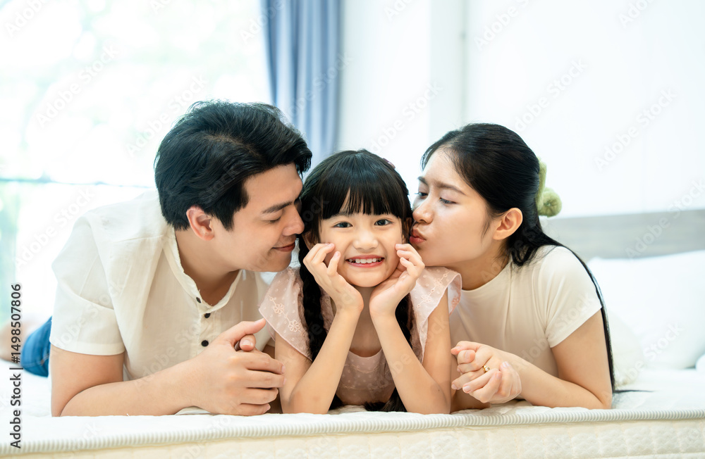 Fototapeta premium A loving family moment where Asian parents kiss their happy daughter on the cheeks while lying on the bed.