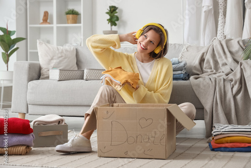 Young woman in headphones packing baby clothes in donation cardboard box on f...