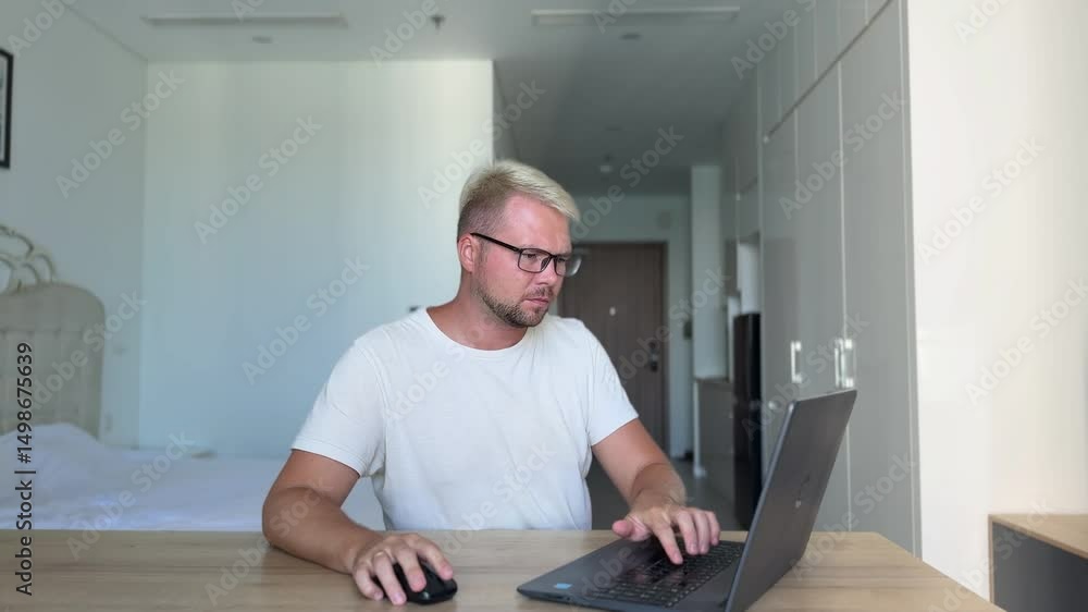 A young guy , blond, in a white t-shirt sits at a desk in a stylish flat and works on his laptop. The laptop breaks down, the man doesn't know what to do and gets angry A young guy , blond, in a white t-shirt sits at a desk in a stylish flat and works on his laptop. The laptop breaks down, the man doesn't know what to do and gets angry