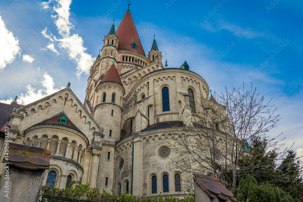 Naklejka premium Stunning view of St. Francis of Assisi Church in Vienna, Austria, showcasing its Romanesque Revival architecture, red rooftops, and majestic towers under a vivid blue sky with scattered clouds.