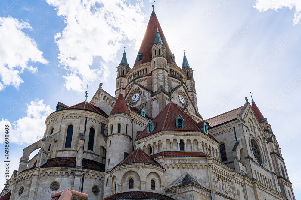 Fototapeta premium Magnificent view of St. Francis of Assisi Church in Vienna, Austria, featuring its iconic Romanesque Revival architecture, clock towers, and red-tiled rooftops against a bright sky.