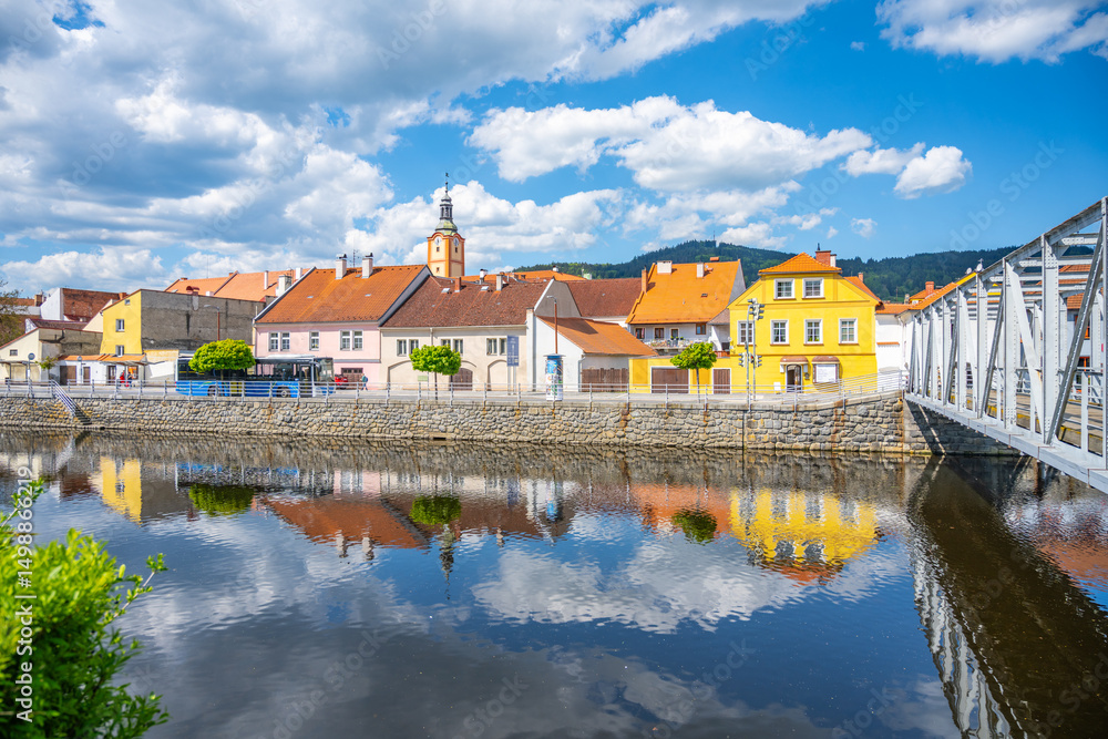 Obraz premium Sunlight reflects off the Otava River in Susice, Czechia, showcasing vibrant townhouses and a quaint bridge against a backdrop of blue skies and fluffy clouds.