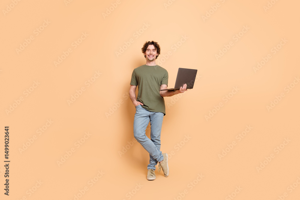 Young man posing casually with a laptop against a beige backdrop, wearing trendy khaki attire Young man posing casually with a laptop against a beige backdrop, wearing trendy khaki attire