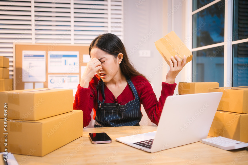 A middle-aged woman, who appears to be Asian, rests her forehead on her hand, looking stressed and disheartened amidst packages and a laptop, possibly due to declining sales. A middle-aged woman, who appears to be Asian, rests her forehead on her hand, looking stressed and disheartened amidst packages and a laptop, possibly due to declining sales.