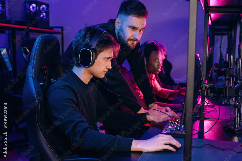 Guy is standing and looking, helping. Gamers are playing video game by computers Guy is standing and looking, helping. Gamers are playing video game by computers