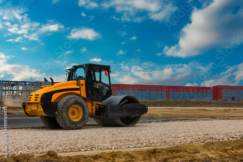 Canvas Print Yellow road roller compactor working on gravel at a construction site with a red and gray industrial building in the background