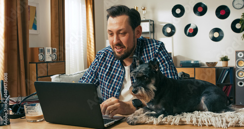 A relaxed brunette man with a beard smiles during a video conference, saying goodbye and showing his domestic dog to the camera, enjoying friendly connection and warmth from home.