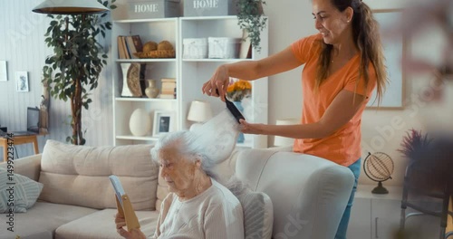 Adult daughter combing her elderly mother's white hair at home in living room.