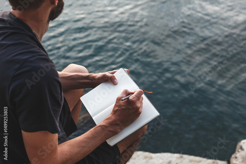 man sits on a rocky shore by water, writing in notebook. he is immersed in his thoughts, journaling peacefully in nature. calm waves, quiet atmosphere. Concept: mindfulness, solitude, mental health.