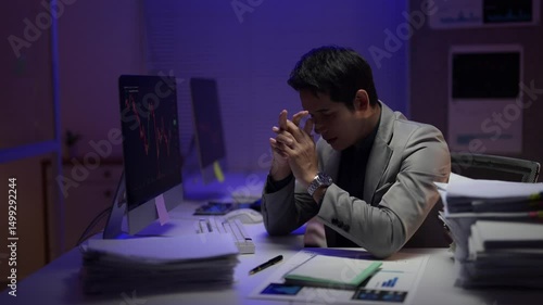 A man is sitting at a desk with a computer monitor in front of him. He is wearing a suit and he is in a state of distress