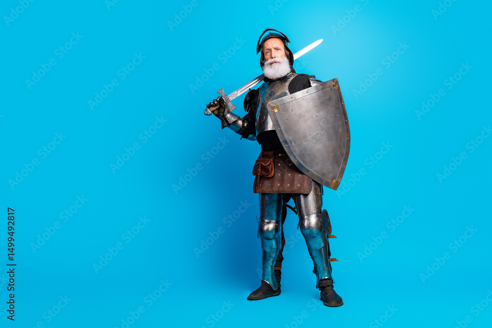 Naklejka premium Elderly man dressed as a knight in medieval armor with sword and shield, standing confidently against a bright blue background