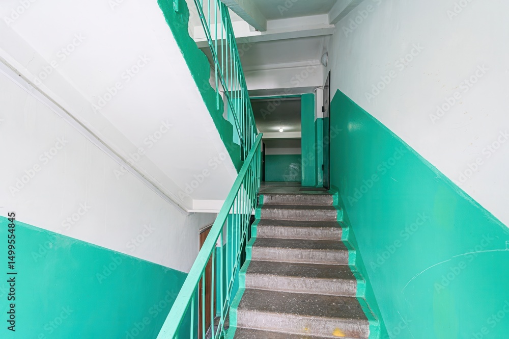 Fototapeta premium Stairwell with teal and white walls. Concrete stairs with teal railing lead upwards. A doorway is visible at the top of the stairs