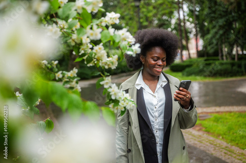 Smiling black businesswoman walking in a city park after rain, using mobile phone app near blooming jasmine bush, enjoying good news and pleasant fragrance