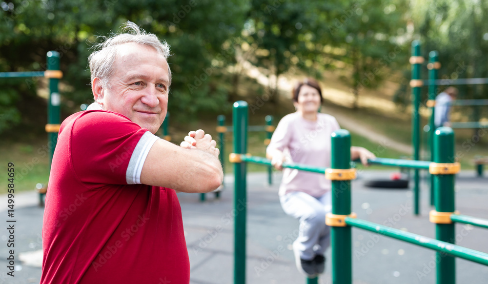 Obraz premium Staying after retirement. Happy joyful mature retired sportsman wearing sportswear doing side stretching exercises with arm over his head, exercising outside in city park