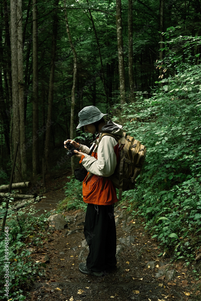 Fototapeta premium Young man hiking through dense green forest with camera in hand, wearing backpack and rain jacket, exploring nature and documenting.