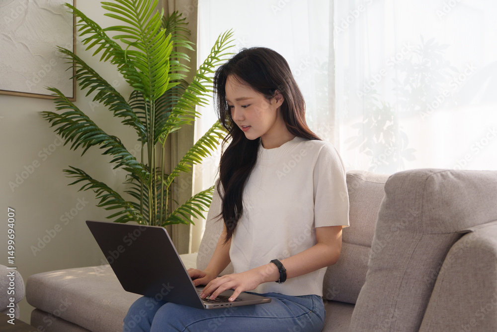 Young asian woman sitting on a sofa, working on a laptop in a bright and cozy living room. Young asian woman sitting on a sofa, working on a laptop in a bright and cozy living room.