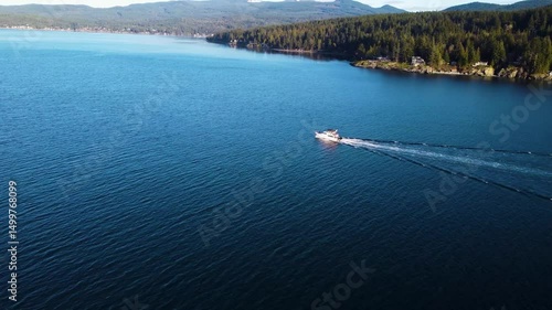 Aerial view of a boat slowly moving across a calm ocean. Close to the tree-filled coastline we cross the boats wake
