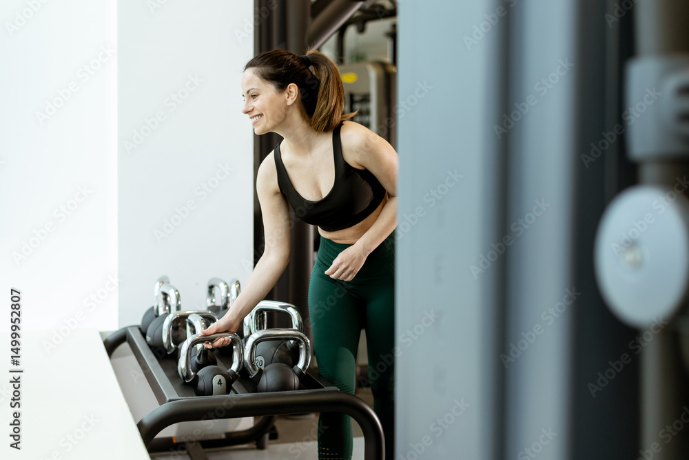 Fototapeta premium Woman enjoys strength training in a modern gym while selecting kettlebells during her fitness journey