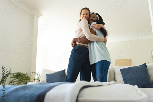 Hugging diverse female friends on white sofa cushions in living room, with pillows and plant