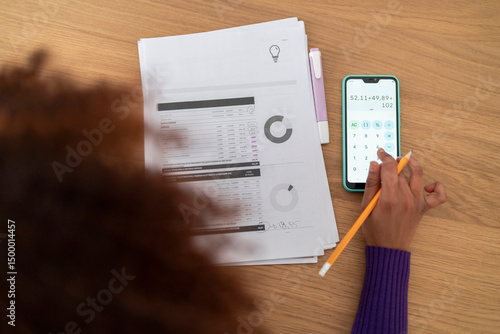 A person uses a phone calculator to check figures on financial documents. They are likely managing expenses or verifying calculations for personal or business purposes.
