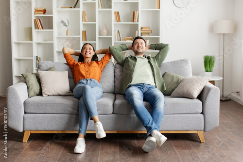 Konstfotografi Happy relaxed couple leaning on couch and holding hands behind head, enjoying comfort of their modern home
