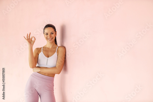 Portrait of fit sportswoman in shape leaning on a wall and gesturing okay at the camera.