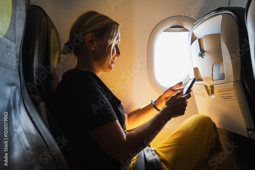 Passenger woman is flying in plane. Girl using phone sitting by airplane window. Traveling female with smartphone inside plane enjoying flight. Woman using wifi connection on board. Traveling girl