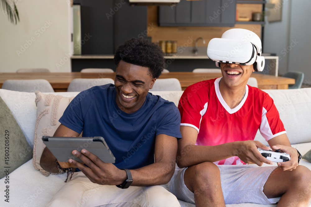 © Wavebreak Media - Laughing Diverse male friends using tablet and VR headset on sofa in open-plan living room