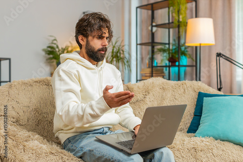 Quarrel. Furious annoyed young Hindu man freelancer engage in heated conversations sitting on sofa couch. Upset Indian guy male expresses frustration using laptop computer on couch in apartment room.